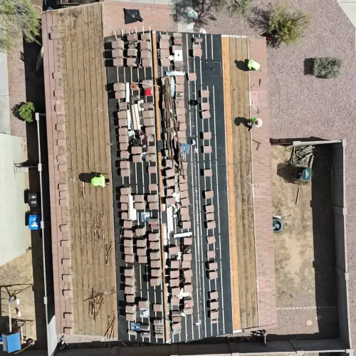 An aerial view of a residential roof under construction, with new reddish-brown shingles laid out in rows and workers visible.