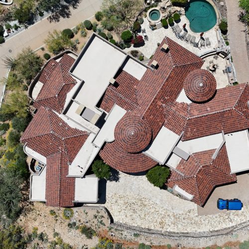 An aerial, top-down view showcases a large, sprawling luxury home with a distinctive reddish-brown tile roof and multiple architectural features. The property includes white flat roof sections, circular towers with conical tile roofs, and extensive landscaping with desert vegetation and paved driveways. A swimming pool and patio area are visible towards the top right of the property. A bright blue vehicle is parked near the front entrance.